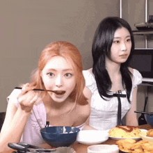 two women are sitting at a table with plates of food and one is eating with chopsticks