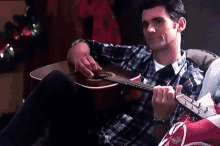 a man in a plaid shirt sits on a couch playing an acoustic guitar