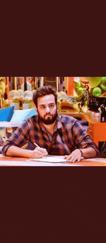 a man in a plaid shirt is sitting at a desk writing on a piece of paper