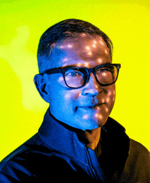 a man wearing glasses and a blue jacket stands in front of a yellow background