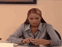 a woman in a gray shirt is sitting at a desk holding a cell phone .