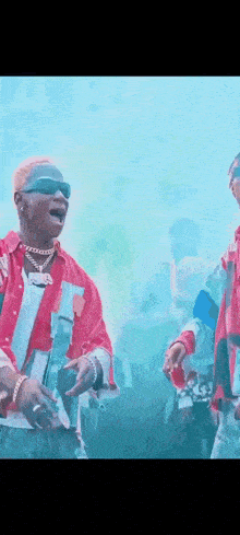 two men are standing next to each other in front of a crowd . one of the men is wearing sunglasses .