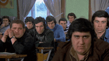 a group of men sit at desks in a classroom with one man looking at the camera