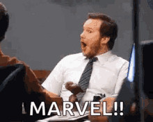 a man in a suit and tie is sitting at a table with his mouth open and says marvel .