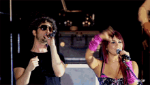 a woman singing into a microphone next to a man wearing sunglasses and a black shirt