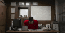 a man in a red shirt sits at a desk with a computer monitor