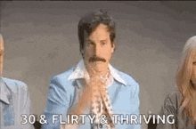 a man with a mustache is sitting in front of a group of people and clapping his hands .