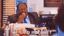 a man in a suit and tie is sitting at a desk and eating a sandwich .
