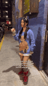 a girl wearing a blue sweater with a teddy bear on it stands in an alleyway