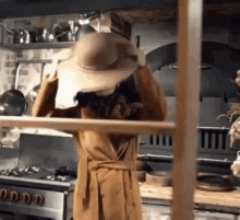 a woman in a brown coat is holding a hat over her head in a kitchen .