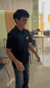 a young man wearing glasses is standing in a classroom with tables and chairs .