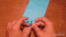 a person is folding a piece of blue paper on a wooden table with the words made in animotica below them
