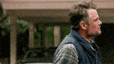 a man wearing a vest and plaid shirt is standing in front of a car .