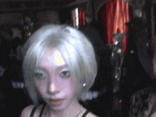 a woman with short white hair looks at the camera with a blurry background