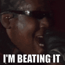 a man wearing sunglasses is singing into a microphone with the words `` i 'm beating it '' written below him .
