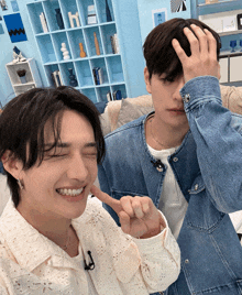a man in a denim jacket holds his hand to his forehead next to another man