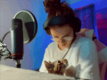 a woman wearing headphones and glasses holds a cat in her lap in front of a microphone