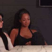 two women are sitting at a table talking to each other . one of the women is wearing glasses .
