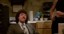 a man with curly hair and glasses is sitting in a kitchen with a bag of gas in the background
