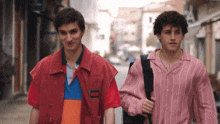two young men walking down a street with one wearing a pink striped shirt and the other wearing a red vest