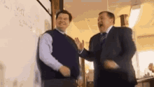 two men in suits are standing next to each other in front of a whiteboard and laughing .