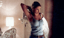 a woman in a blue tank top and jeans is standing in a bedroom