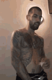 a shirtless man with a beard and tattoos is standing in front of a wall .