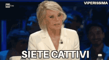 a woman in a white suit and glasses is sitting at a table and saying siete cattivi