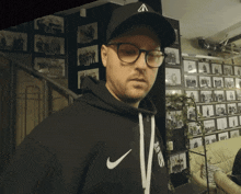 a man wearing glasses and a black nike hoodie stands in front of a wall of pictures