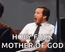 a man in a suit and tie is sitting on a couch with his mouth open and says `` holy fuck mother of god '' .