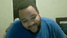 a man with a beard and a blue shirt is smiling while sitting on a bed .
