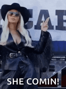 a woman wearing a cowboy hat and sunglasses is standing in front of a sign that says `` she comin ! ''