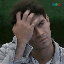 a man in a denim jacket scratches his head with his hand