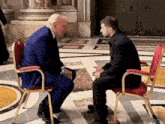 a man in a suit sits in a red chair talking to another man in a black suit