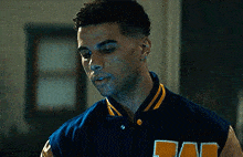 a young man wearing a blue and yellow varsity jacket is standing in a room .