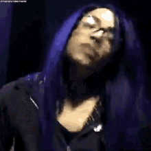 a woman with purple hair and glasses is wearing a black jacket and making a funny face .