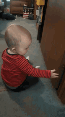 a baby in a red and black striped shirt is sitting on the floor reaching for a door handle