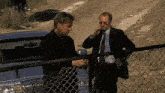 two men are standing next to a chain link fence talking on their cell phones