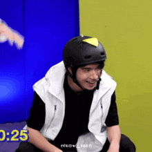 a man wearing a black helmet and a white vest is smiling while sitting in front of a blue wall ..
