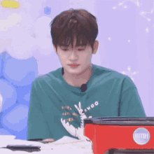 a young man in a green shirt is sitting at a table with a red pot on it .