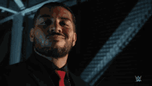 a man with a beard wearing a black suit and red tie with a w logo in the background