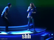 a woman singing into a microphone while a man stands behind her and says shh on the bottom