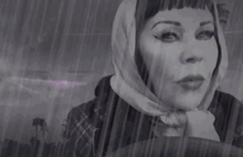 a black and white photo of a woman wearing a scarf driving a car in the rain .