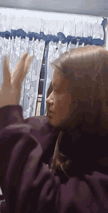 a woman in a purple sweater is standing in front of a window with curtains