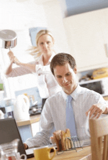a man sitting at a table with a laptop and a woman holding a pot in the background