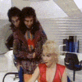 a woman in a red top is sitting in a chair while two men stand behind her