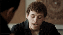 a man with curly hair is talking to another man and the words jayanedna are on the bottom right