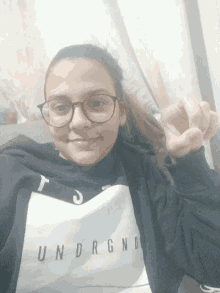a girl wearing glasses and a shirt that says underneath is giving the peace sign