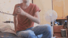 a man in a red shirt is sitting on a bed with a fan in the background