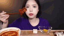 a woman in a purple shirt is eating a piece of food with chopsticks